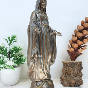 May include: A bronze statue of the Virgin Mary, standing on a base with a snake, wearing a crown and a blue robe. The statue is on a white surface.
