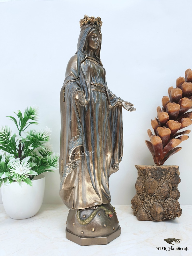 May include: A bronze statue of the Virgin Mary, standing on a base with a snake, wearing a crown and a blue robe. The statue is on a white surface.