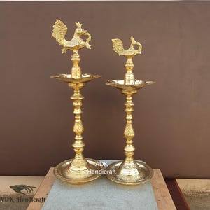 May include: Two ornate, gold-colored brass lamps. Each lamp features a detailed peacock design at the top, a central oil dish, and a tall, decorative stem. The lamps are standing on a round base. The text "ADK Handicraft" is visible.