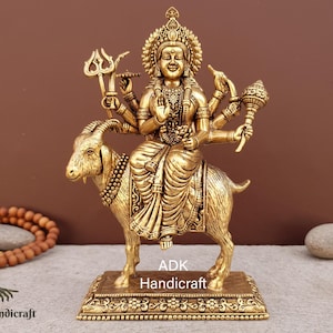 Puede incluir: Una estatua de metal dorado de una diosa hindú con múltiples brazos montando una cabra. La diosa lleva una túnica fluida y una corona. La estatua está sobre una base con detalles intrincados. "ADK Handicraft" está escrito en la base.