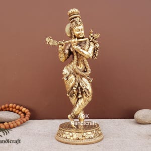 Puede incluir: Una estatua dorada de una deidad tocando la flauta, con detalles intrincados en la ropa y las joyas. La estatua se encuentra sobre una base decorativa, con un pavo real en el hombro. El fondo es de color marrón liso.