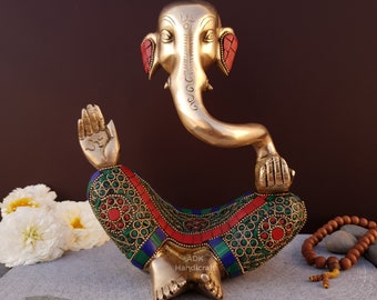May include: A gold-colored brass statue of Ganesha, the Hindu god of wisdom and prosperity, sitting in a meditative pose. The statue is decorated with colorful inlaid stones, including red, green, and blue. The statue is sitting on a brown surface.