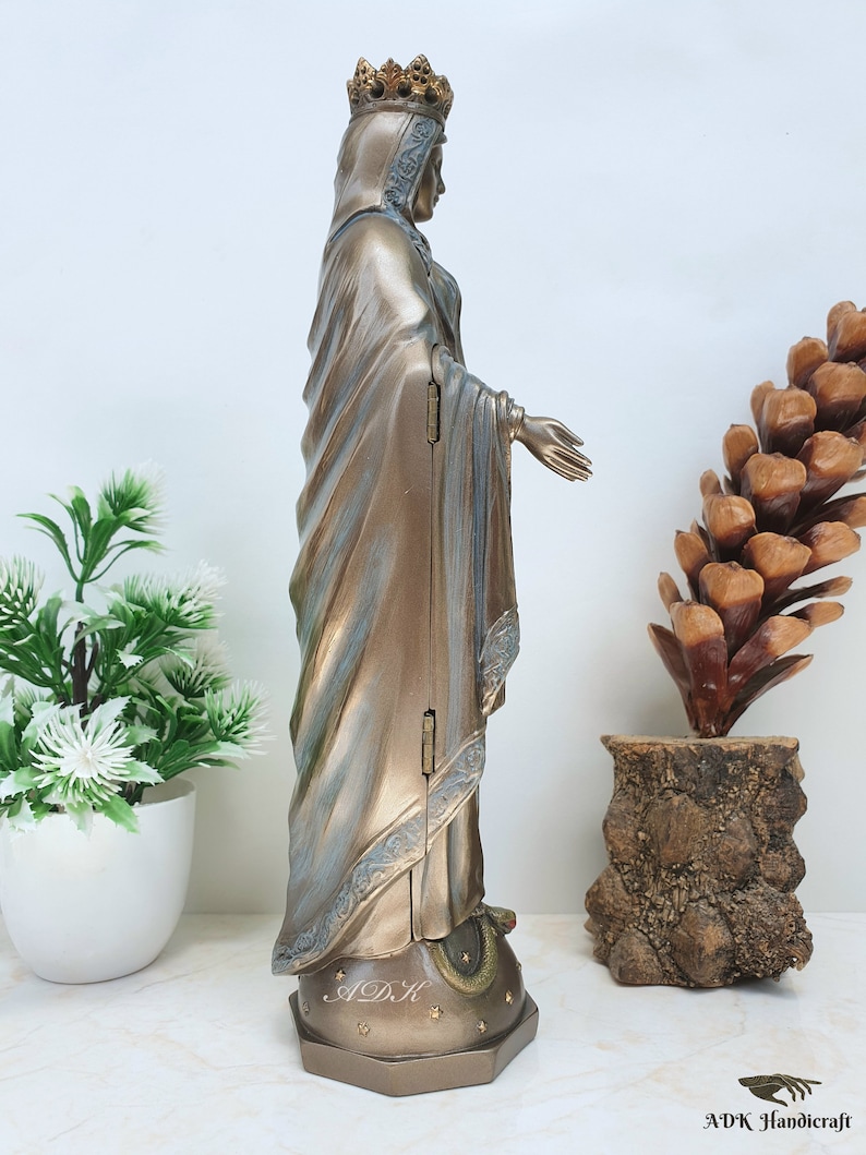 May include: A gold-toned statue of the Virgin Mary, standing on a star-shaped base with the letters "ADK" inscribed. The statue is wearing a crown and a flowing robe.
