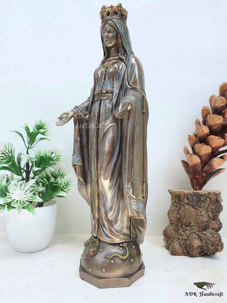 May include: A bronze statue of the Virgin Mary standing on a base with a snake and stars. The statue is wearing a blue robe and a crown. The statue is about 12 inches tall.