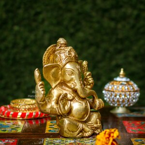 May include: A gold-colored brass statue of Ganesha, the Hindu god of beginnings, wisdom, and prosperity. The statue is sitting on a colorful wooden table with a flower in the foreground.