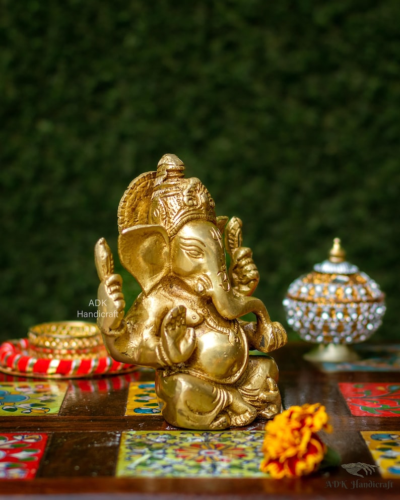 May include: A gold-colored brass statue of Ganesha, the Hindu god of beginnings, wisdom, and prosperity. The statue is sitting on a colorful wooden table with a flower in the foreground.