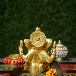 May include: A gold-colored metal statue of Ganesha, the Hindu god of wisdom and prosperity, sitting with his hands raised in a gesture of blessing. The statue is on a colorful tiled surface with a yellow flower in the foreground.