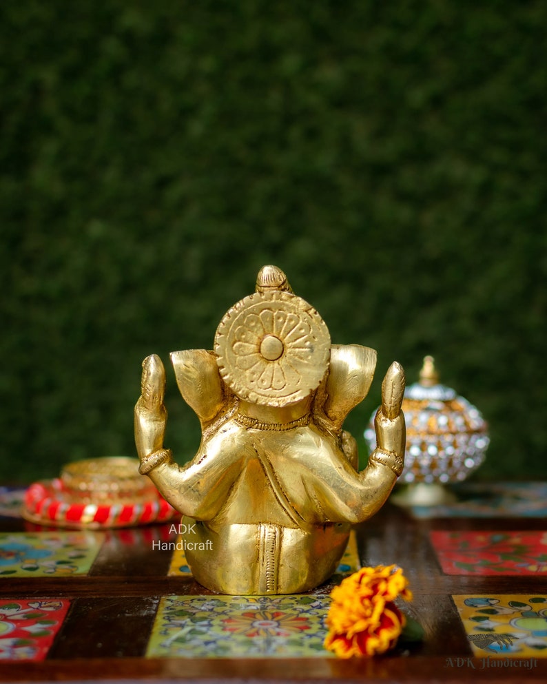 May include: A gold-colored metal statue of Ganesha, the Hindu god of wisdom and prosperity, sitting with his hands raised in a gesture of blessing. The statue is on a colorful tiled surface with a yellow flower in the foreground.