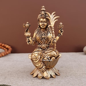 Puede incluir: Una estatua dorada de una deidad sentada, adornada con intrincadas joyas y una corona. La figura está sentada sobre una base de flor de loto y sostiene objetos en ambas manos. La estatua tiene un diseño detallado y probablemente está hecha de latón.