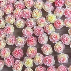 May include: A collection of miniature rose flower embellishments. The roses are in shades of pink and yellow, with a gradient effect. A black circular tag with the number 35 in white is also present. The roses are scattered on a light brown wooden surface.