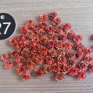 May include: A collection of small, red and gold-toned rose-shaped embellishments scattered on a wooden surface. A black circular tag with the number 27 in white is in the upper left corner. These decorative elements are ideal for crafts.