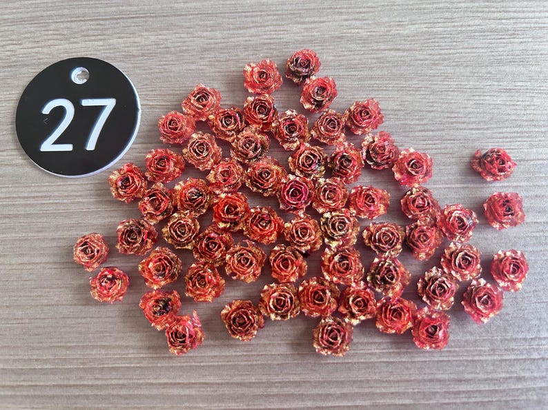 May include: A collection of small, red and gold-toned rose-shaped embellishments scattered on a wooden surface. A black circular tag with the number 27 in white is in the upper left corner. These decorative elements are ideal for crafts.