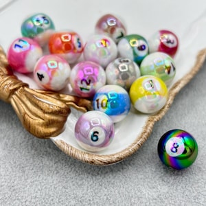 Puede incluir: Una colección de bolas de billar iridiscentes con fondos blancos y marcas numeradas, exhibidas en un plato decorativo blanco y dorado. Las bolas son de varios colores, incluyendo rosa, azul y amarillo. Una bola es de un vibrante color arcoíris.