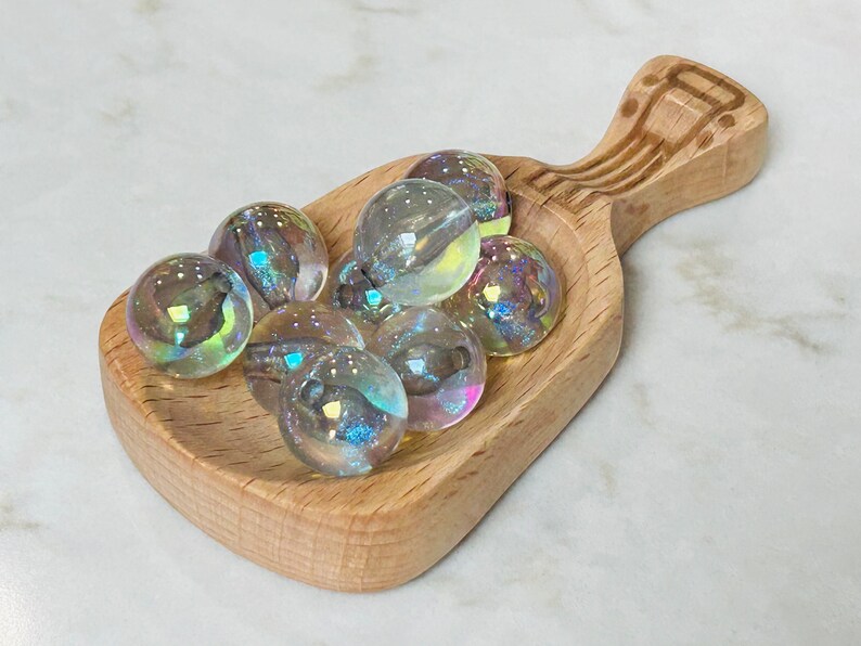 May include: A wooden scoop filled with iridescent glass marbles. The marbles are various shades of blue, green, and purple.