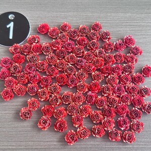 May include: A collection of small, red and gold-edged artificial roses scattered on a gray surface. A black circular tag with the number "1" in white is in the upper left corner. These decorative roses are ideal for crafts.