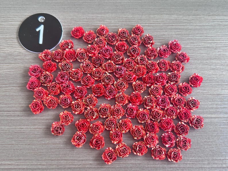 May include: A collection of small, red and gold-edged artificial roses scattered on a gray surface. A black circular tag with the number "1" in white is in the upper left corner. These decorative roses are ideal for crafts.