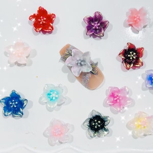 May include: An assortment of colorful, flower-shaped nail art decorations. The decorations are in various colors, including red, purple, pink, blue, and yellow. One nail is decorated with a flower and glitter. The decorations are scattered on a white surface.