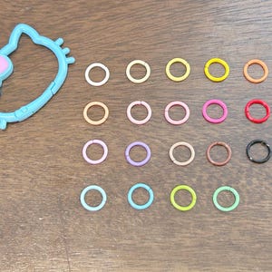 Puede incluir: Un dije de plástico azul claro con forma de cabeza de gato con un lazo rosa, junto a una colección de pequeños anillos metálicos de colores. Los anillos son de varios colores, incluyendo rosa, amarillo y verde.