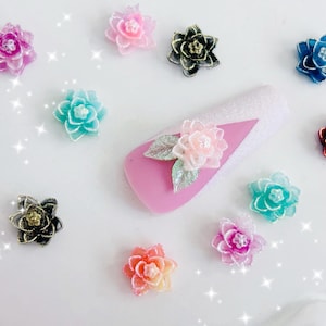 May include: A collection of colorful flower-shaped nail art decorations. The assortment includes various shades of pink, blue, green, and red, with some featuring glitter accents. A pink nail with a flower design is also present.