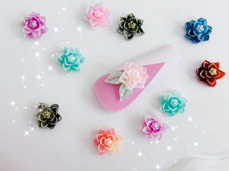 May include: A collection of small, multi-colored 3D flower charms in shades of pink, blue, orange, black, and teal, featuring glittery centers. One pink flower with silver leaves is attached to a pink and white glittery nail art display tip.