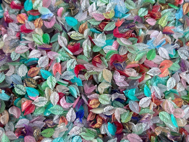 May include: A large collection of small, leaf-shaped beads in a variety of translucent and opaque colors including pink, green, blue, red, and gold. Each bead features a detailed, embossed vein pattern on the surface, creating a textured appearance.