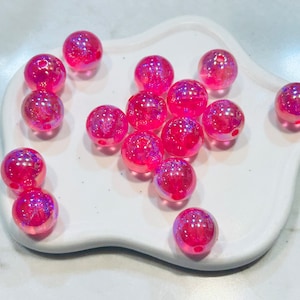 May include: A group of pink iridescent glass beads with a hole in the centre. The beads are scattered on a white surface.