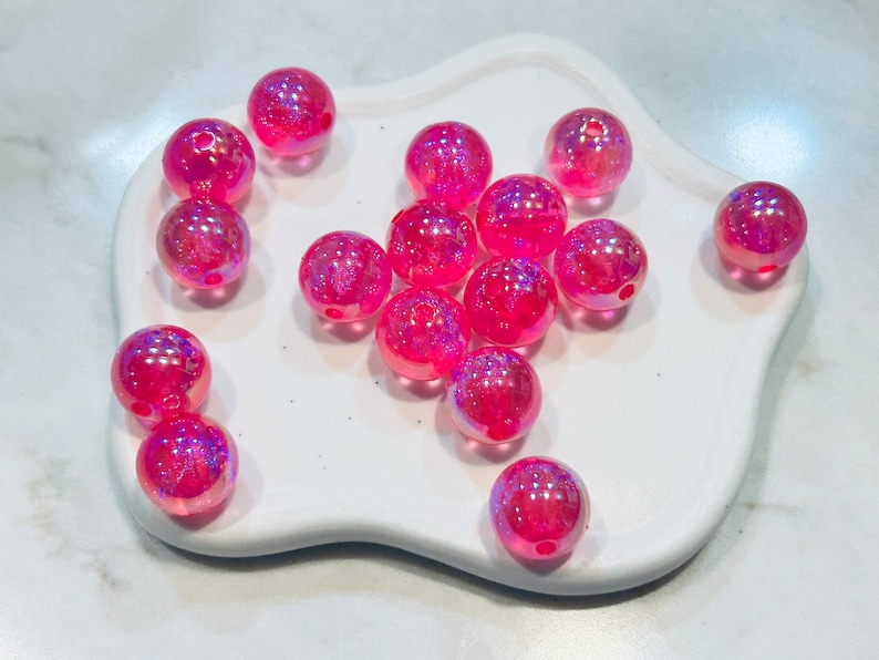 May include: A group of pink iridescent glass beads with a hole in the centre. The beads are scattered on a white surface.
