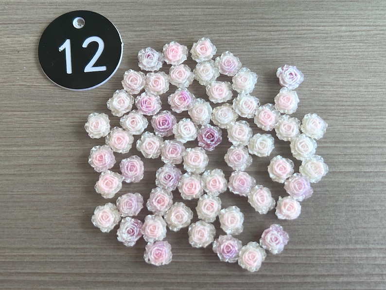 May include: A collection of small, translucent rose-shaped embellishments in shades of pink and white. A black circular tag with the number 12 in white is in the upper left corner. These decorative flowers are ideal for crafts.
