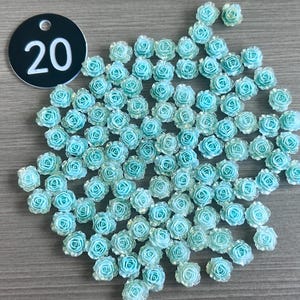 May include: A collection of small, light blue rose-shaped embellishments. The roses are clustered together, with a few scattered around. A black tag with the number "20" in white is in the upper left corner. These decorative elements are suitable for crafts and DIY projects.