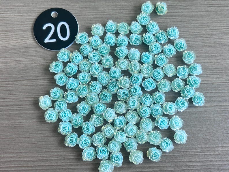 May include: A collection of small, light blue rose-shaped embellishments. The roses are clustered together, with a few scattered around. A black tag with the number "20" in white is in the upper left corner. These decorative elements are suitable for crafts and DIY projects.