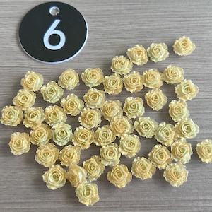 May include: A collection of small, yellow-toned artificial rose flowers scattered on a light brown surface. A black circular tag with the number "6" in white is also present. These decorative flowers are ideal for crafts.