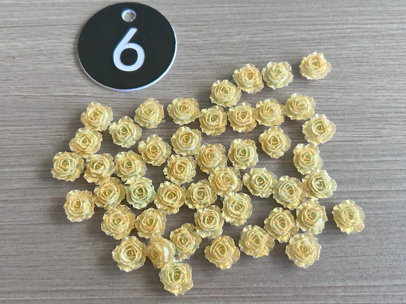 May include: A collection of small, yellow-toned artificial rose flowers scattered on a light brown surface. A black circular tag with the number "6" in white is also present. These decorative flowers are ideal for crafts.