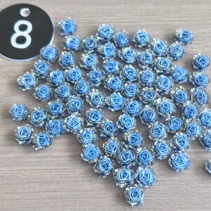 May include: A collection of small, blue and silver rose-shaped embellishments. A black tag with the number "8" in white is in the upper left corner. The roses are scattered on a light wood-grain surface.