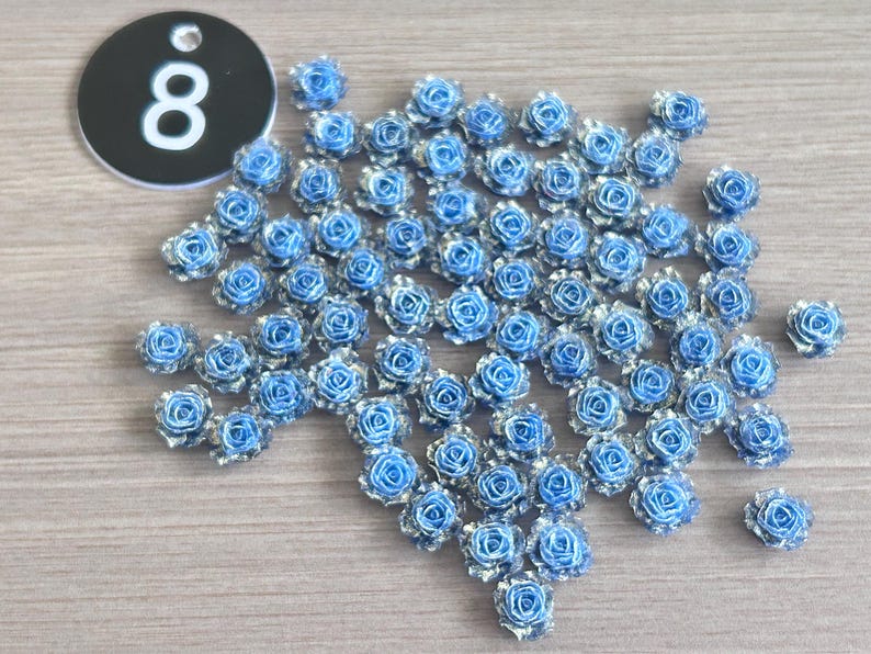 May include: A collection of small, blue and silver rose-shaped embellishments. A black tag with the number "8" in white is in the upper left corner. The roses are scattered on a light wood-grain surface.