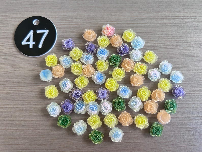 May include: A collection of small, colorful rose-shaped embellishments in various pastel shades, including yellow, blue, purple, green, and peach. A black tag with the number 47 is in the upper left corner. These are likely craft supplies.