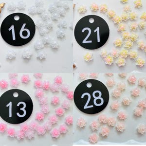 May include: A collection of small, multi-petaled flower cabochons with central beads, organized into four groups labeled with black circular tags numbered 16, 21, 13, and 28. The flowers appear in white, yellow-pink, light pink, and peach tones.