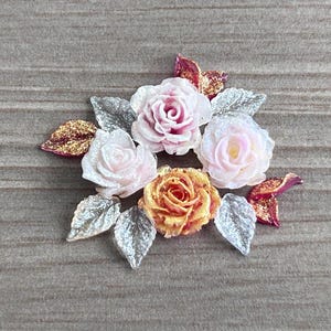 May include: A decorative floral embellishment featuring three roses in shades of soft pink, pale white, and vibrant orange. The arrangement is accented with several silver-toned leaves and smaller, glittery magenta-colored petals.