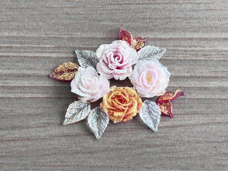 May include: A decorative floral embellishment featuring three roses in shades of soft pink, pale white, and vibrant orange. The arrangement is accented with several silver-toned leaves and smaller, glittery magenta-colored petals.