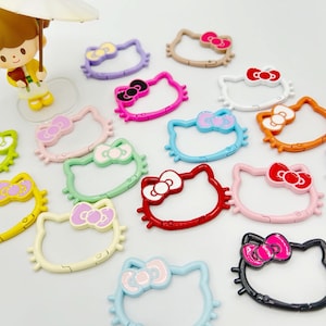 May include: A collection of colorful Hello Kitty-shaped keychains. The keychains feature a variety of colors, including pink, red, blue, green, and yellow, with a bow detail. A figurine of a girl holding an umbrella is in the background.