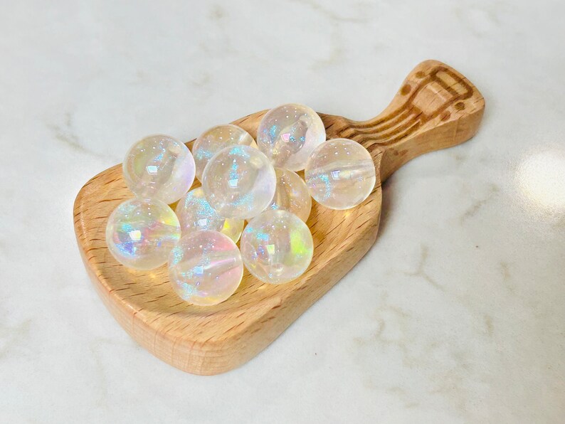 May include: A wooden scoop filled with iridescent white glass beads. The beads are round and have a rainbow sheen.