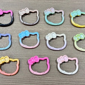 May include: A collection of colorful Hello Kitty-shaped keychains. Each keychain features a bow and a gradient color scheme, including pink, blue, green, and yellow. The keychains are arranged on a neutral surface.