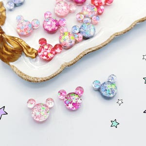 May include: A collection of glittery, mouse-shaped embellishments in shades of pink, red, and blue, are scattered on a white surface. Some are arranged on a decorative dish with gold accents. Star doodles decorate the image.