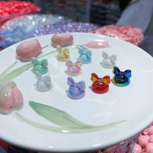 Pu&ograve; includere: Un piatto bianco mostra perline colorate a forma di fiocco e perline rosa a forma di tulipano. I fiocchi sono in vari colori, tra cui iridescente, rosso, giallo, blu e verde. Le perline sono probabilmente per la creazione di gioielli o artigianato.