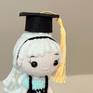 May include: A handmade crochet doll with white hair, wearing a blue dress and a black graduation cap with a yellow tassel. The doll has black button eyes and a light skin tone. The doll is a charming, handcrafted item.