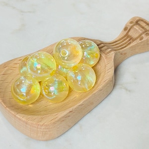May include: A wooden scoop filled with iridescent yellow glass beads. The beads are round and have a slight shimmer to them.