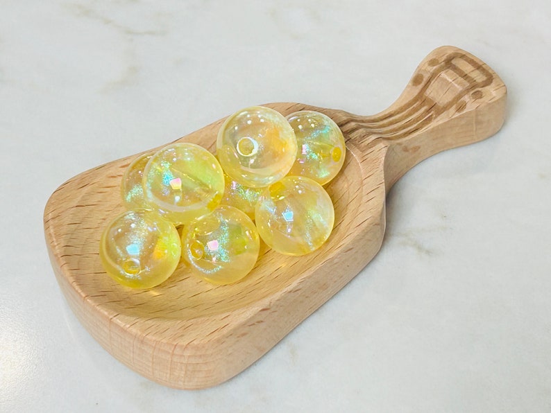 May include: A wooden scoop filled with iridescent yellow glass beads. The beads are round and have a slight shimmer to them.