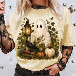May include: A pale yellow t-shirt featuring a graphic print of a white ghost holding a lit lantern, a black crow perched on a stack of books, a small orange pumpkin, and a larger white pumpkin, all surrounded by green ivy leaves and vines.