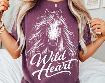 May include: A plum-coloured t-shirt with a white graphic of a horse's head and flowing mane. The words "Wild Heart" are written in a cursive font below the horse image. The shirt is a crew neck style.