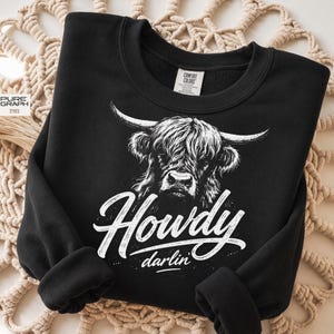 May include: Black sweatshirt with a white Highland cow graphic and the words "Howdy darlin'". The sweatshirt has a crew neck and long sleeves. The Comfort Colors label is visible. The design is a rustic, country style.