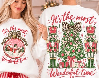 May include: A white sweatshirt featuring a graphic of a Christmas ornament with a snowman and houses inside, and a back graphic of a Christmas tree with two nutcrackers, gifts, and a leopard print bow, with the text It's the most wonderful time.